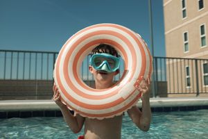 Boy wearing goggles holds striped inflatable pool ring at outdoor swimming pool on sunny day.