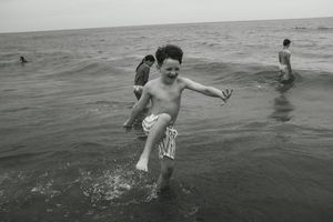 Black and white photo of a joyful boy running through ocean waves at the beach with other children.