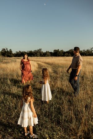 Family of four in a golden field at sunset, two girls in white dresses facing parents under a clear blue sky.