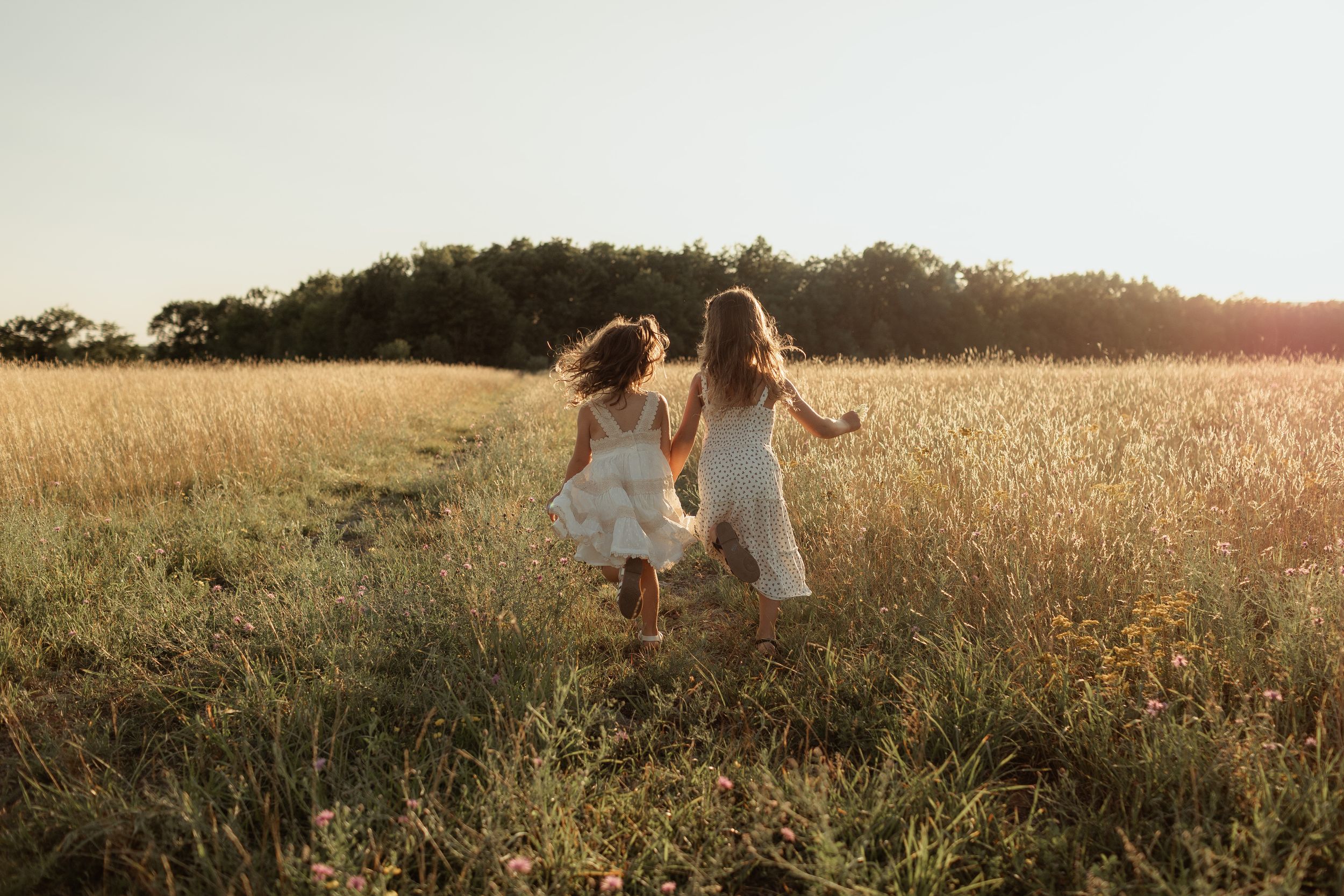 Two young girls in white dresses run hand-in-hand through a golden field at sunset.