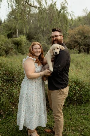 A smiling couple holds a white Labrador puppy outdoors surrounded by lush green trees and foliage.