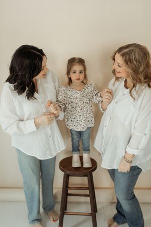 A small child stands on a wooden stool between two women in white shirts and jeans in a minimalist setting.