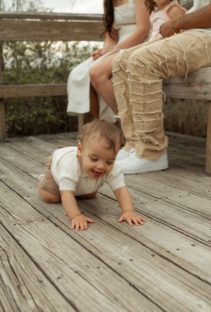 A toddler in beige pants crawls on weathered wooden deck boards while being watched by an adult.