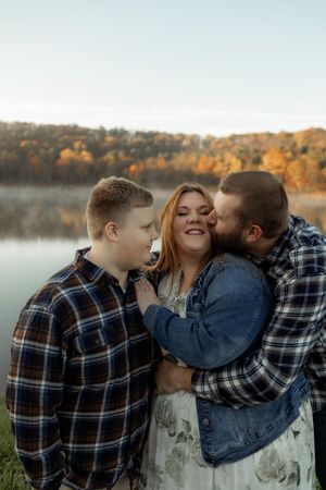 Family of three sharing a tender moment by an autumn lake, with parents embracing and smiling son nearby.