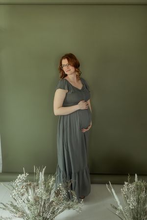 A person in a gray dress stands against a sage green wall with dried white floral arrangements displayed below.