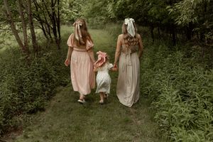 Two people in flowing dresses walk hand in hand through a lush green forest setting during golden hour.