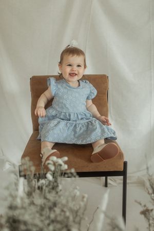 A smiling baby in a light blue lace dress sits on a wooden chair against a white backdrop with delicate flowers.
