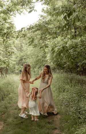 Three figures in soft pastel dresses gather in a lush green garden setting on a summer afternoon.