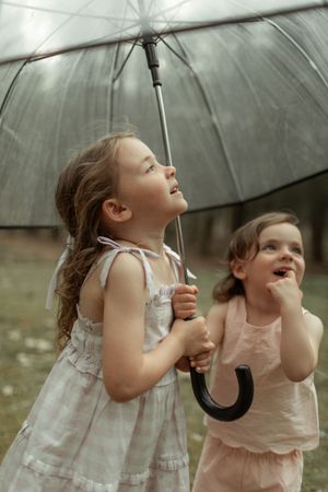 Two young children in light dresses look up while holding clear umbrellas outdoors.