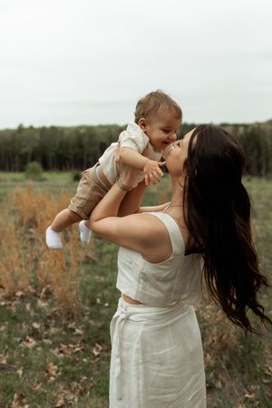 A tender moment captured in a field as a parent lifts and plays with their baby in a white dress against a natural backdrop.