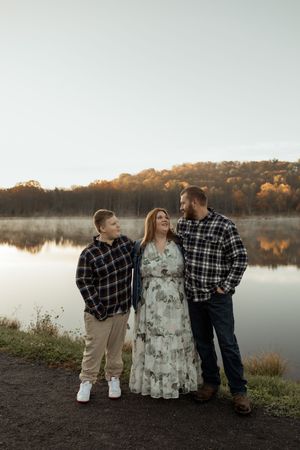 Family of three posing by a misty lake at sunset, surrounded by autumn foliage in a warm outdoor portrait.
