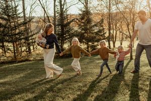 Family of six holding hands and running through a sunlit autumn park with tall trees and golden grass.