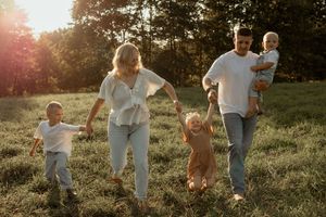 Happy family walking in sunny green field, parents holding hands with toddlers, golden hour light, outdoor family photo.