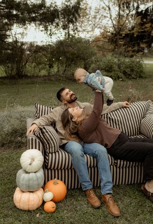 A couple sits on a striped outdoor sofa with pumpkins, lifting their baby in the air during a fall family photo session.