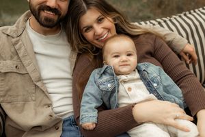 Happy young family with smiling parents and adorable baby in denim jacket posing together on cozy outdoor seating.