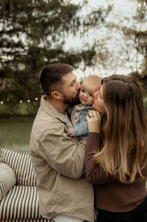 A young couple kisses their smiling baby outdoors near a pond with string lights and lush trees.