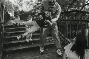 Black and white photo of a father playfully lifting two young children on wooden deck steps while a woman watches nearby.