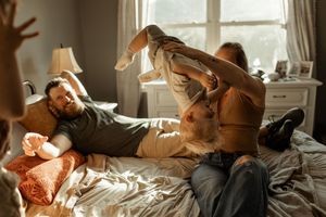 Two adults playfully lift a baby on a sunlit bed, creating a joyful family moment at home.