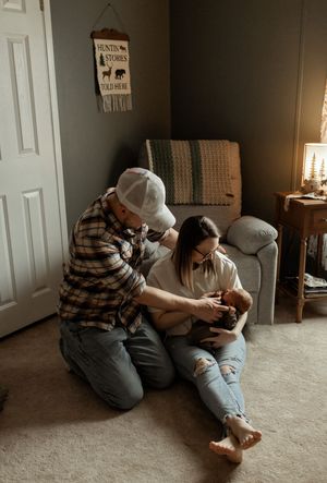 A couple sits on the floor together, lovingly cradling their newborn baby in a cozy, warmly lit nursery room.