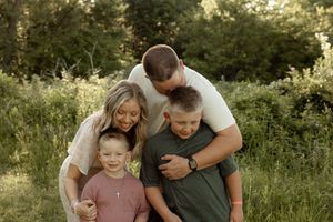 Family of four hugging outdoors in a lush green field, parents embracing two young boys in a warm portrait.