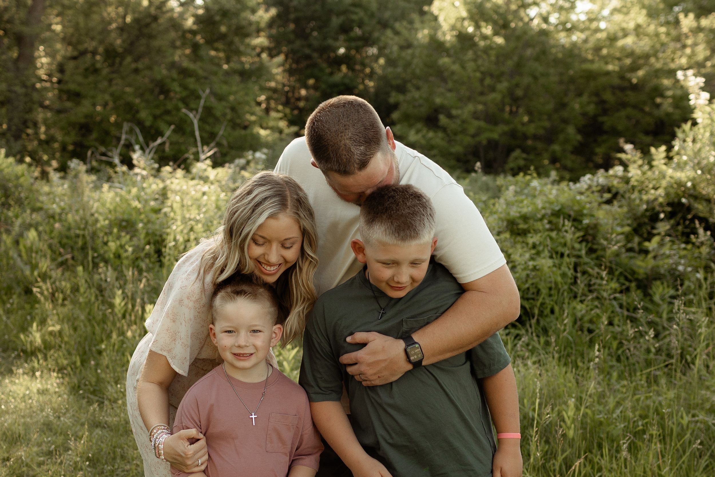 Family of four hugging outdoors in a lush green field, parents embracing two young boys in a warm portrait.