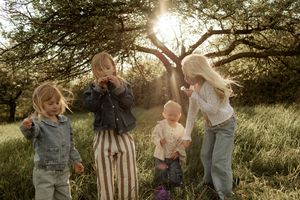 Four young children playing outdoors in a sunlit meadow beneath a large tree on a warm sunny day.