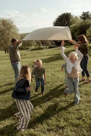 Family playing outdoors on green grass, lifting a white sheet while children laugh and run beneath it on a sunny day.