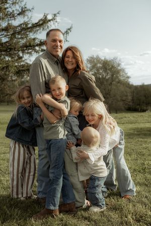 A happy family of six poses outdoors on green grass, with two parents hugging four young blonde children near trees.