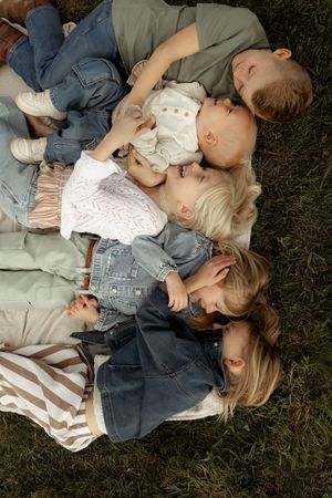 A family of five lies together on grass, with parents and three young children in denim outfits enjoying a peaceful outdoor moment.