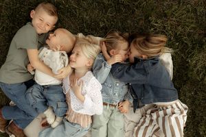 Five young children lying together on grass, cuddling and kissing in a sweet family portrait outdoors.