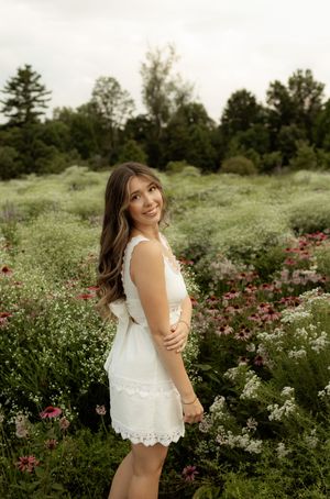 Young woman in white lace dress smiling in a lush wildflower field with pink and white blooms.