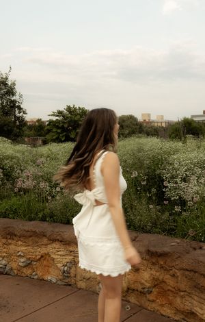 A young woman in a white cutout dress twirls outdoors near a stone wall with wildflowers and greenery behind her.