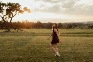Young woman in black dress twirling on green lawn at golden sunset with trees and hills in background.