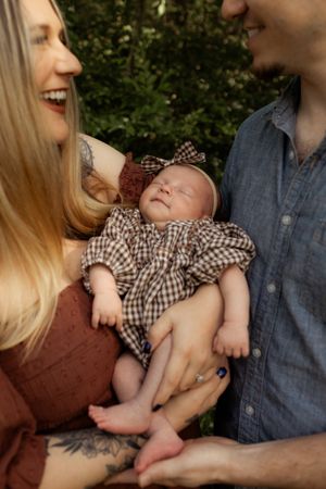 Happy parents smiling and holding their newborn baby girl in a plaid outfit outdoors.