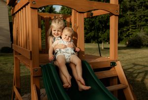 Two young children sitting together on a green backyard playground slide on a sunny day.
