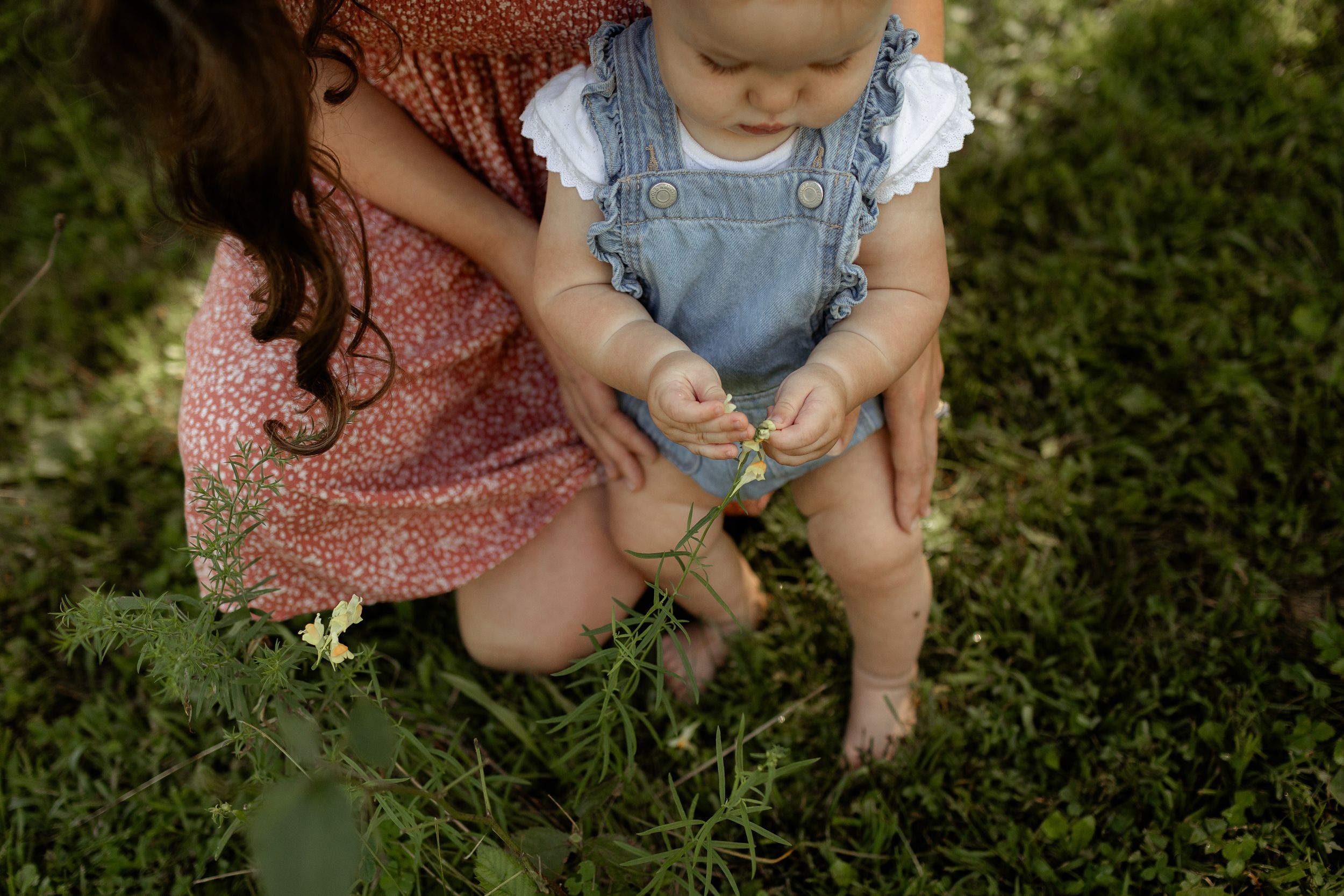 Mother kneeling on grass helping baby in denim overalls explore nature outdoors.