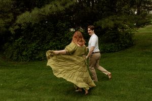 Couple dancing joyfully outdoors, woman in flowing green dress with red hair, man in white shirt and tan pants, lush garden.
