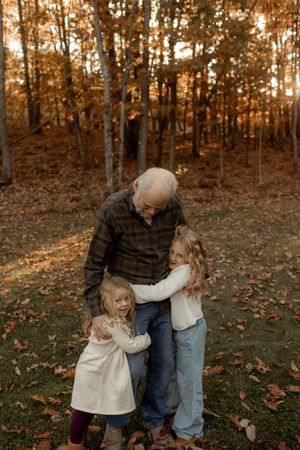 Grandfather hugging two young granddaughters outdoors in autumn forest with golden fall foliage.