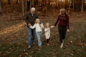 Family of four walking hand-in-hand through autumn woods with fallen leaves, two young girls between grandpa and mom.