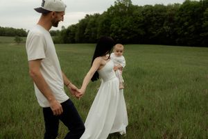 Young family with baby walks hand-in-hand through lush green field, mother in white dress holding infant.