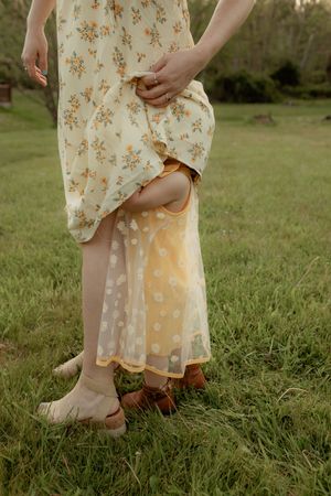 A young child hiding behind a woman in a floral dress, both standing on green grass outdoors.