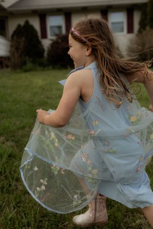 A young girl twirls in a light blue floral tulle dress outdoors, her hair flowing as she spins joyfully in a yard.