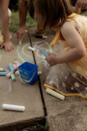 Young girl in yellow daisy dress drawing with blue chalk on sidewalk, with colorful chalk pieces and blue bucket nearby.