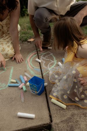 Family drawing colorful chalk art on concrete patio, with a blue bucket and chalk pieces scattered nearby.