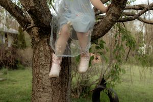 Child in a light blue dress and pink boots sitting in the branches of a tree in a garden.