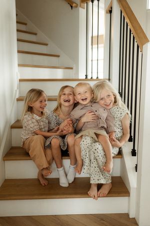 Four young children laughing and hugging together on wooden staircase steps in a bright, modern home.