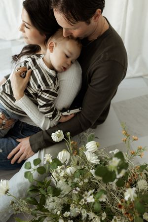 A loving family of three with parents cradling a sleepy toddler surrounded by elegant white floral arrangements.