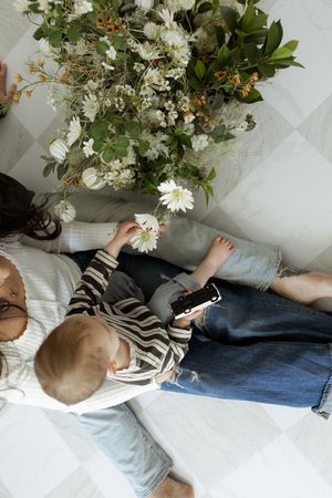 Overhead view of mother and child sitting on floor with white floral arrangement, child holding phone and flower.