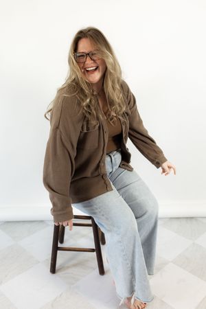 Smiling woman with glasses wearing brown cardigan and light jeans sitting on wooden stool against white background.