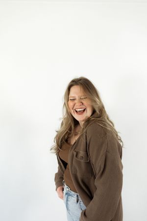 Laughing woman with long blonde hair wearing brown ribbed cardigan and light jeans against white background.